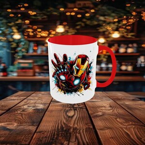 May include: A white ceramic mug with a red interior and handle. The mug features a colorful illustration of Iron Man in a dynamic pose, with red, gold, and blue accents. The design is set against a backdrop of paint splatters.