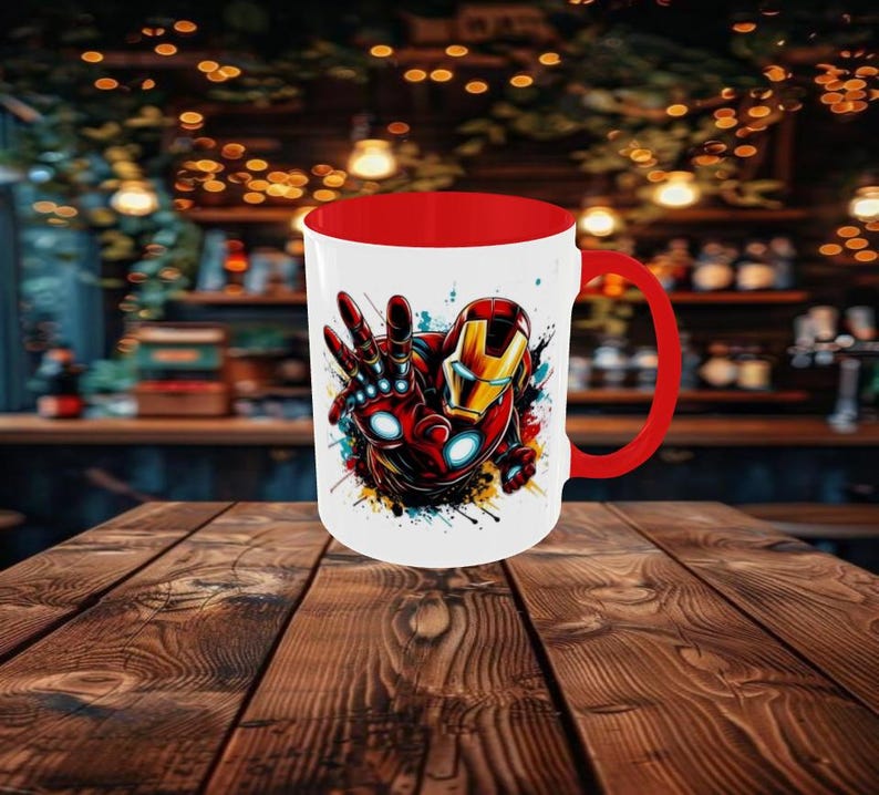 May include: A white ceramic mug with a red interior and handle. The mug features a colorful illustration of Iron Man in a dynamic pose, with red, gold, and blue accents. The design is set against a backdrop of paint splatters.