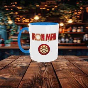 May include: White ceramic mug with a blue interior and handle. The mug features the words "IRON MAN" in red and gold lettering, along with a circular emblem with a red and gold design. The mug is set on a wooden table.