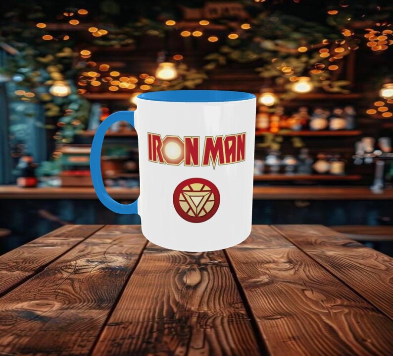 May include: White ceramic mug with a blue interior and handle. The mug features the words "IRON MAN" in red and gold lettering, along with a circular emblem with a red and gold design. The mug is set on a wooden table.