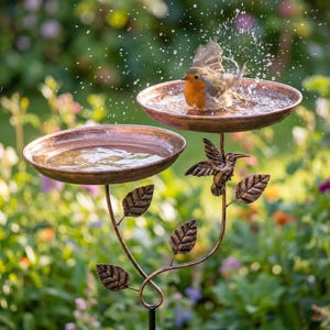 May include: A bronze-coloured, two-tiered bird bath with a small bird splashing in the upper basin. The bird bath features a decorative hummingbird and leaf design. The background is a blurred garden scene with green foliage and colourful flowers.