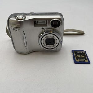 May include: Silver Nikon Coolpix 4100 digital camera with a zoom lens and flash. Includes a blue and gold PNY Optima SD card. The camera has a wrist strap attached.
