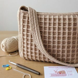 May include: A beige crocheted bag with a waffle stitch pattern, featuring a long strap. A ball of yarn, a crochet hook, yarn needle, and stitch markers are next to the bag. The text "THE BEIGE WAFFLE BAG" is visible on the bag.