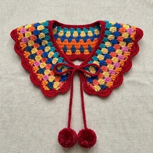 May include: A crochet collar featuring a multi-colored granny square pattern in shades of blue, yellow, orange, and pink. The collar has a solid red scalloped border and includes two long red ties that end in matching red yarn pom-poms.