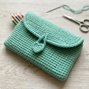Puede incluir: Un estuche de ganchillo verde menta con cierre de solapa y botón en forma de corazón. El estuche está lleno de lápices de colores. Tijeras y una aguja con hilo en el fondo.