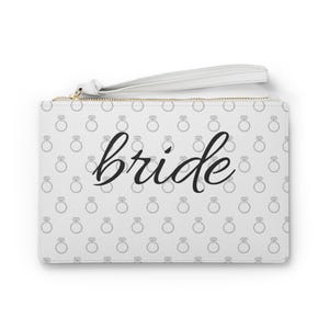 Puede incluir: Clutch blanca con cremallera dorada y correa de muñeca. El bolso está estampado con pequeñas ilustraciones de anillos de diamantes grises y la palabra "bride" en escritura negra. Un accesorio de boda.