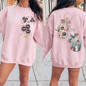 May include: Pale pink sweatshirt with a fantasy-themed design. The front has a group of black, fluffy characters with big eyes, with stars and leaves. The back features two potion bottles with floral and forest illustrations.