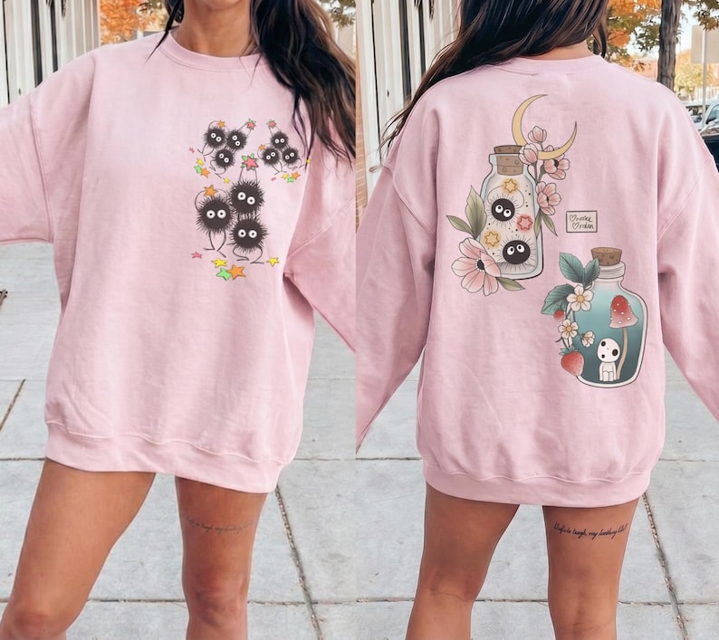May include: Pale pink sweatshirt with a fantasy-themed design. The front has a group of black, fluffy characters with big eyes, with stars and leaves. The back features two potion bottles with floral and forest illustrations.