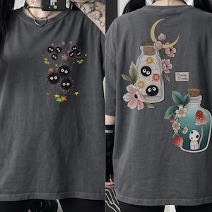 May include: Charcoal grey t-shirt with a front graphic of black, fluffy creatures with large eyes and colourful stars. The back of the shirt features two illustrated jars with floral and moon designs, and a small heart with text.