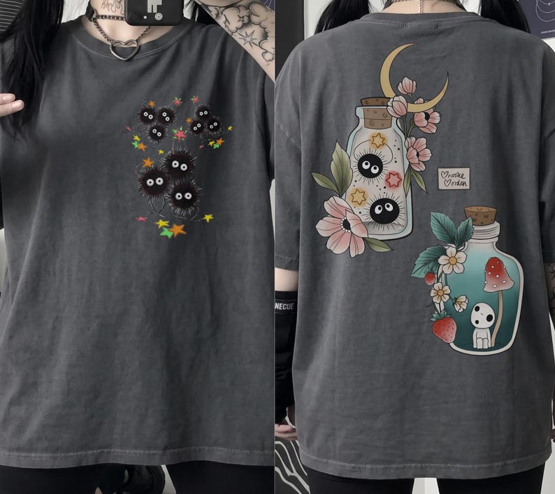 May include: Charcoal grey t-shirt with a front graphic of black, fluffy creatures with large eyes and colourful stars. The back of the shirt features two illustrated jars with floral and moon designs, and a small heart with text.