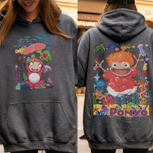 K&ouml;nnte beinhalten: Dunkelgrauer Kapuzenpullover mit einer farbenfrohen Cartoon-Illustration des Films "Ponyo" auf Vorder- und R&uuml;ckseite. Die Vorderseite zeigt die Figur unter einem roten Regenschirm, die R&uuml;ckseite zeigt die Figur mit dem Wort "Ponyo".