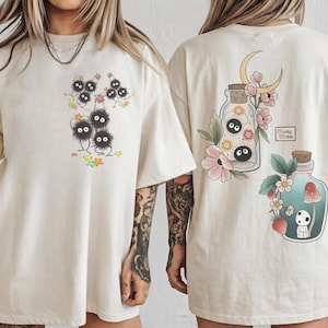 May include: Cream-coloured t-shirts with whimsical illustrations. One shirt shows a group of black, fluffy creatures with star accents. The other shirt showcases a jar with a moon and flowers, and a second jar with a mushroom and a small creature.