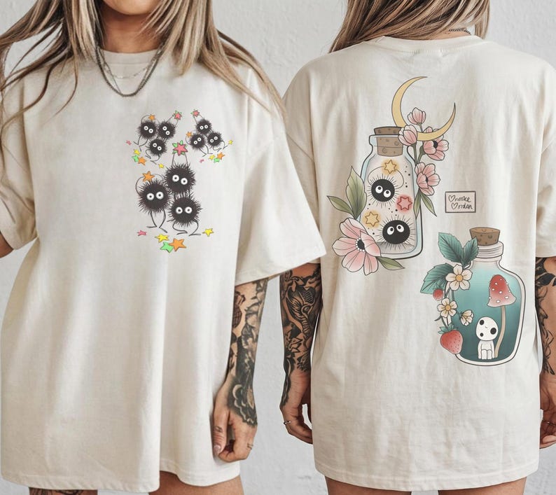 May include: Cream-coloured t-shirts with whimsical illustrations. One shirt shows a group of black, fluffy creatures with star accents. The other shirt showcases a jar with a moon and flowers, and a second jar with a mushroom and a small creature.