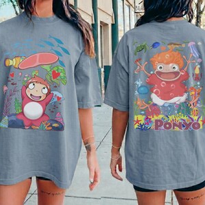 K&ouml;nnte beinhalten: Ein blau-graues T-Shirt mit einem Cartoon-Motiv der Figur "Ponyo" aus dem Animationsfilm. Das Frontdesign zeigt die Figur mit Meeresbewohnern. Das R&uuml;ckendesign zeigt die Figur mit dem Wort "Ponyo".