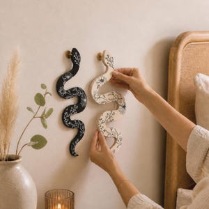 May include: Two decorative snake-shaped wall hangings. One is black with white floral detailing, the other is natural wood with black floral detailing. Both have gold-coloured knobs for hanging. The image shows the natural wood snake being held up.