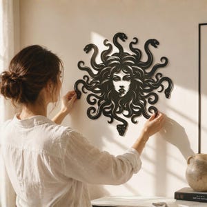 Può includere: Un'opera d'arte murale nera di Medusa con dettagli intricati. L'opera presenta un volto centrale con capelli e serpenti fluenti. I serpenti sono disposti attorno al volto, creando un design sorprendente e dettagliato. Il pezzo è montato su un muro bianco.