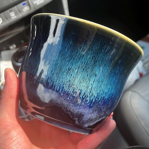 Puede incluir: Una taza de cerámica con un esmalte reactivo que presenta una base azul marino profundo que transiciona hacia un patrón de goteo en turquesa y azul claro. El borde tiene una banda fina de color amarillo pálido y una textura brillante.