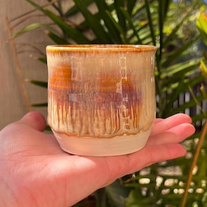May include: A small ceramic cup with a cylindrical shape, featuring a glossy glaze that transitions from a creamy beige at the top to a rich, deep amber and brown drip pattern, ending in a solid, unglazed off-white base at the bottom.