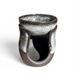May include: A ceramic bowl with a speckled interior sits atop a dark brown, U-shaped holder. The holder has a glossy, light blue and brown glaze on the edges. The piece is designed for serving or display.