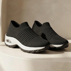 May include: A pair of black slip-on sneakers featuring a textured knit upper with horizontal ribbed detailing, a thick white cushioned midsole, and a visible air pocket unit integrated into the heel section of the sole for support.
