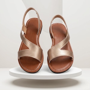 May include: A pair of flat sandals featuring metallic gold straps that cross over the top of the foot and a slingback heel strap. The footbed is brown with visible white stitching along the edges and a black sole base for a simple design.
