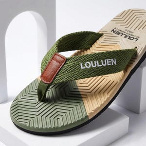 May include: Close-up of a green and tan flip-flop. The flip-flop has a green strap with the word "LOULUEM" in white. The sole is tan and green with a geometric pattern. A brown leather accent is on the strap.