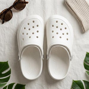 May include: A pair of white clogs with multiple ventilation holes, next to tortoiseshell sunglasses and a pair of beige socks. Green monstera leaves are placed near the clogs on a white linen surface. Casual footwear.