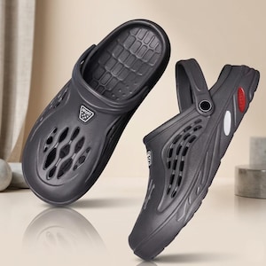 May include: Two dark gray clogs with ventilation holes and a strap. The clogs have a textured footbed and a small logo on the side. One clog is angled, showing the side and the back strap.