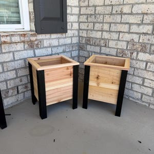 May include: Two square planters constructed from natural light-coloured wood panels with a smooth finish. Each planter features four vertical black corner posts that extend to form legs and a matching light wood trim around the top square opening.