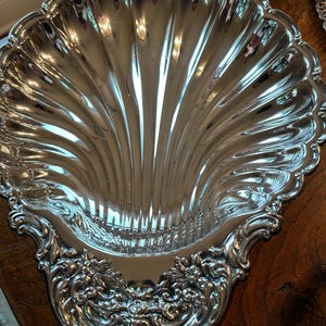 May include: A silver-colored, shell-shaped serving dish with a fluted interior and ornate floral and scrollwork detailing. The dish has a scalloped edge and reflects the surrounding environment. The decorative piece is likely vintage or antique.