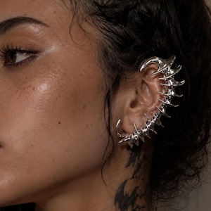 May include: A silver-toned ear cuff featuring a segmented, spine-like structure that curves along the outer edge of the ear. The metallic piece has a polished, reflective finish with multiple pointed protrusions extending from the central curved spine.