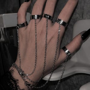 Könnte beinhalten: Eine Hand mit mehreren silbernen Ringen, die durch zarte Ketten verbunden sind. Die Ringe sind schlichte Bänder, und die Ketten führen zu einem Armband. Die Nägel sind lang und schwarz lackiert, was eine gotische Ästhetik erzeugt.