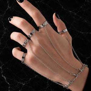 Könnte beinhalten: Ein silberfarbenes Handgeschmeide bestehend aus vier glatten, polierten Metallringen, die durch feine, zarte Gliederketten verbunden sind, welche von jedem Finger zu einem passenden Kettenarmband führen und ein strukturiertes Schmuckstück bilden.