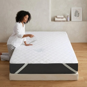 May include: A white mattress protector with a diamond-quilted top surface and dark gray sides featuring light gray corner straps. The protector is positioned on a mattress, demonstrating its water-resistant properties as liquid is poured onto it.
