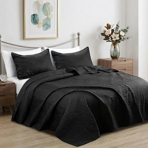 May include: A solid black quilt set featuring a repeating circular stitched pattern. The set includes two matching rectangular pillow shams and a large bedspread that drapes over the mattress, providing a uniform dark aesthetic for the bed.