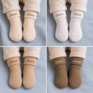 Peut inclure: Quatre paires de chaussettes en tricot côtelé de couleur crème, blanche, beige et marron, chacune dotée d'un revers replié brodé du nom James dans une police cursive marron. Les chaussettes sont portées sur un pantalon clair.