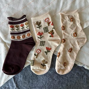 May include: Three pairs of crew socks. The first is dark brown with horizontal stripes and small floral motifs. The second is cream with a scattered pattern of red and brown flowers. The third is beige with a textured diamond pattern and small brown accents.