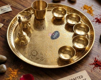 Puede incluir: Un juego de puja de latón que incluye una bandeja circular grande con grabados florales, una campana, una taza pequeña y seis cuencos. Una pegatina en la bandeja dice MKRK y un letrero de madera dice HANDCRAFTED BRASS PUJA SET.