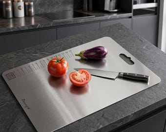 Puede incluir: Una tabla de cortar rectangular de acero inoxidable con acabado cepillado, que incluye una regla impresa en el borde superior, un orificio para colgar y la marca Panagia. Contiene un tomate entero, una rodaja de tomate, una berenjena y un cuchillo.