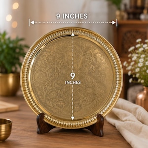 Puede incluir: Una bandeja redonda de metal dorado con un intrincado patr&oacute;n floral grabado en su superficie. La bandeja tiene un borde estriado decorativo y se exhibe sobre un soporte de madera oscura. El texto en la imagen indica 22,86 cm de di&aacute;metro.