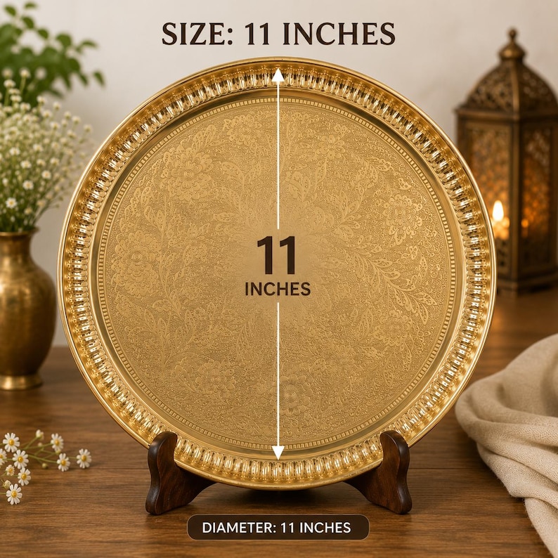 Puede incluir: Una bandeja redonda de metal color dorado con un intrincado patr&oacute;n floral en relieve en toda su superficie y un borde estriado decorativo. La bandeja se exhibe sobre un soporte de madera oscura e incluye texto que indica un di&aacute;metro de 27,94 cm.
