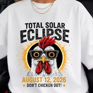 Puede incluir: Una camiseta blanca de manga corta con un gráfico de un pollo con gafas de sol con reflejos de eclipse. El texto dice TOTAL SOLAR ECLIPSE, AUGUST 12, 2026, y DON'T CHICKEN OUT! con un fondo de rayos de sol amarillos tras el pollo.