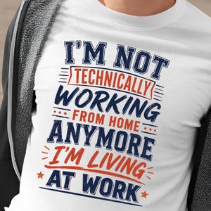 Puede incluir: Una camiseta blanca con texto en azul oscuro y naranja que dice: I'M NOT TECHNICALLY WORKING FROM HOME ANYMORE I'M LIVING AT WORK. El diseño incluye líneas decorativas y estrellas, debajo de una sudadera gris oscuro con cremallera.