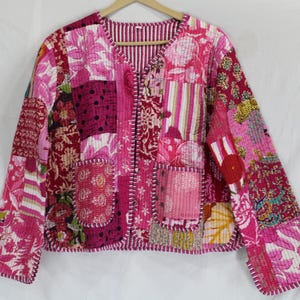 May include: A patchwork jacket in shades of pink, red, and white, featuring floral and geometric patterns. The jacket has a V-neck, two pockets, and a striped lining. The jacket is quilted and has a relaxed fit.