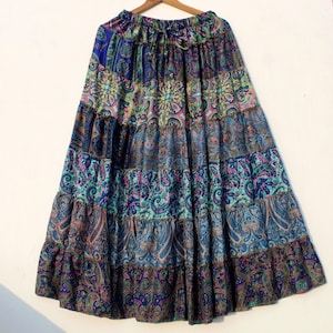 May include: A tiered maxi skirt with a paisley pattern in shades of blue, teal, purple, and brown. The skirt is hanging on a wooden hanger against a white background. The skirt has a drawstring waist.
