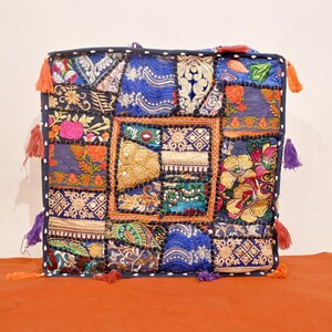 May include: Square patchwork cushion with a navy blue border and colorful embroidered patches. The cushion features various patterns, including floral and geometric designs, with orange, pink, and blue tassels on the corners. The cushion is approximately 18 inches square.