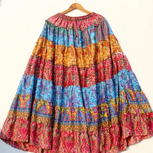 Op de afbeelding: Een gelaagde, zwierige rok met een levendig patchwork-ontwerp. De rok heeft paisley- en bloempatronen in roze, blauw, oranje en geel. Hij hangt aan een houten hanger, wat de volle, uitlopende vorm laat zien.