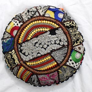 Può includere: Cuscino rotondo patchwork con un vivace mix di colori e motivi. Il cuscino presenta un design centrale ricamato in argento, circondato da un bordo di perline rosse, gialle e bianche. Le sezioni esterne sono composte da varie toppe di tessuto.