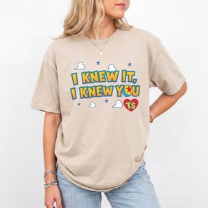 K&ouml;nnte beinhalten: Ein beiges Kurzarm-T-Shirt mit dem Text I KNEW IT, I KNEW YOU und einem roten Herz mit den Buchstaben TS. Das Design enth&auml;lt blaue Sterne und wei&szlig;e Wolkenumrisse, wobei der Buchstabe O in YOU durch einen gelb-blauen Ball ersetzt wurde.