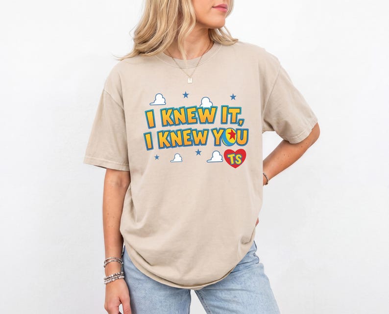 K&ouml;nnte beinhalten: Ein beiges Kurzarm-T-Shirt mit dem Text I KNEW IT, I KNEW YOU und einem roten Herz mit den Buchstaben TS. Das Design enth&auml;lt blaue Sterne und wei&szlig;e Wolkenumrisse, wobei der Buchstabe O in YOU durch einen gelb-blauen Ball ersetzt wurde.